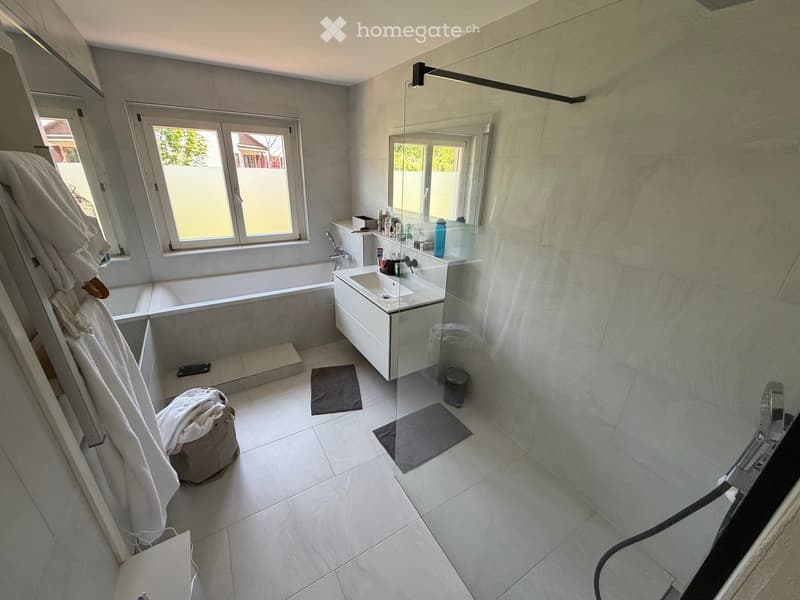 Modern bathroom with a bathtub, sink, mirror, and various toiletries. Features tiled walls and floor, a window, and towels on the side.