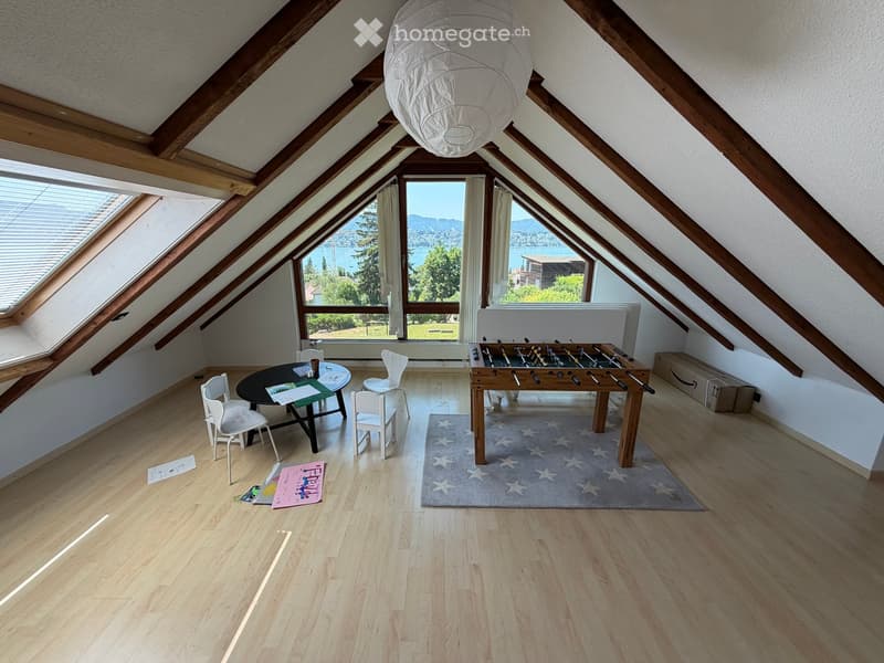Bright attic room with wooden beams, large windows offering scenic views, a round table with chairs, and a football table.