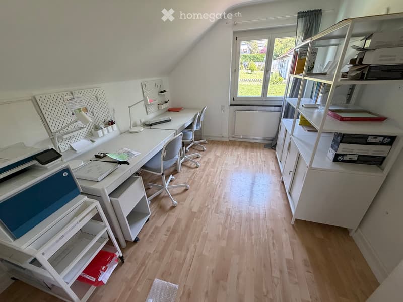 Well-lit attic room with wooden flooring, large window, desk with chair, printer, shelves, and a lamp.