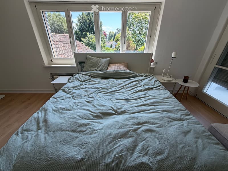 Cozy bedroom with a large bed, nightstand, lamp, and a small stool near the window. The room has wooden flooring and large windows offering a view of the garden.