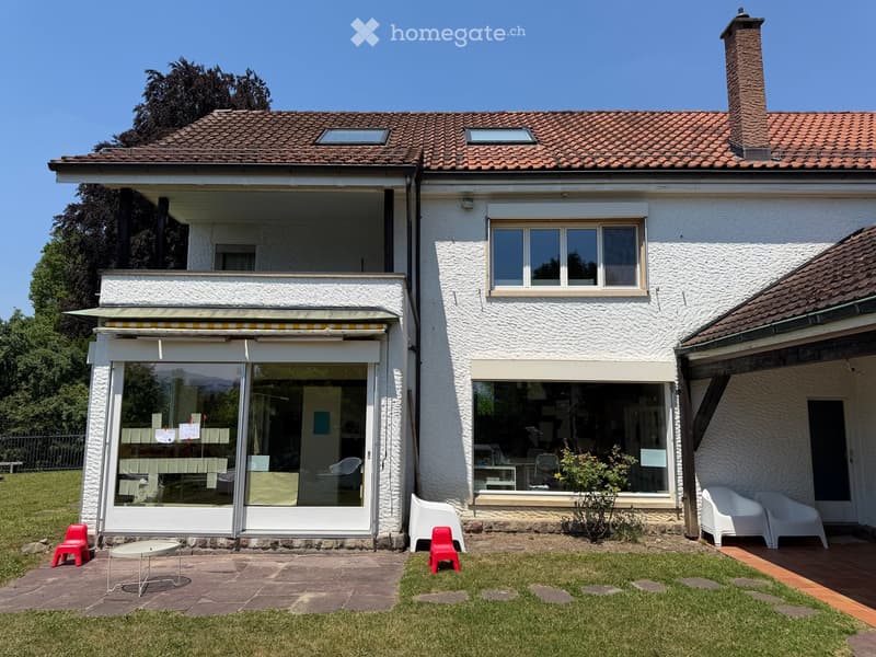 Two-story house with white exterior, large windows, and a balcony. The house features a tiled roof, a chimney, and a covered patio area with glass doors. The surrounding area includes a well-maintained lawn, patio furniture, and a small garden.