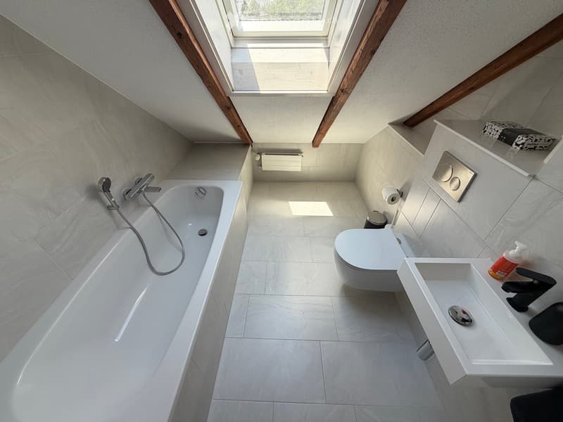 Modern bathroom with a bathtub, toilet, sink, and sleek fixtures. The space features light-colored tiles and wooden beams on the ceiling.