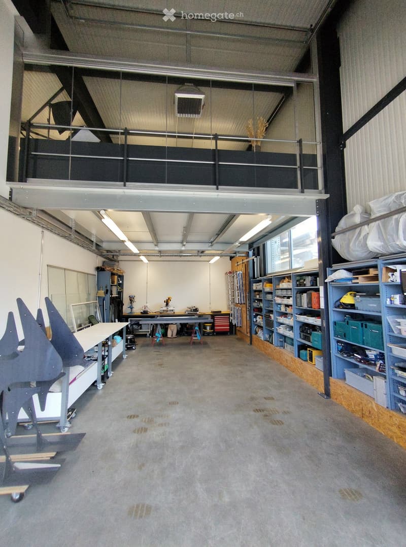 The image shows a spacious, industrial-style workshop or studio with concrete floors, exposed metal beams, and large windows. Visible objects include tables, shelves, stools, and cabinets.
