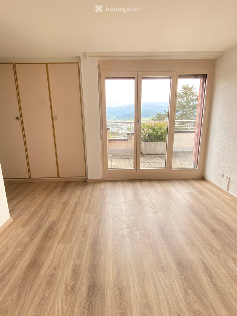 Empty room with light wooden flooring, large sliding glass doors leading to a balcony with a view of mountains and trees.