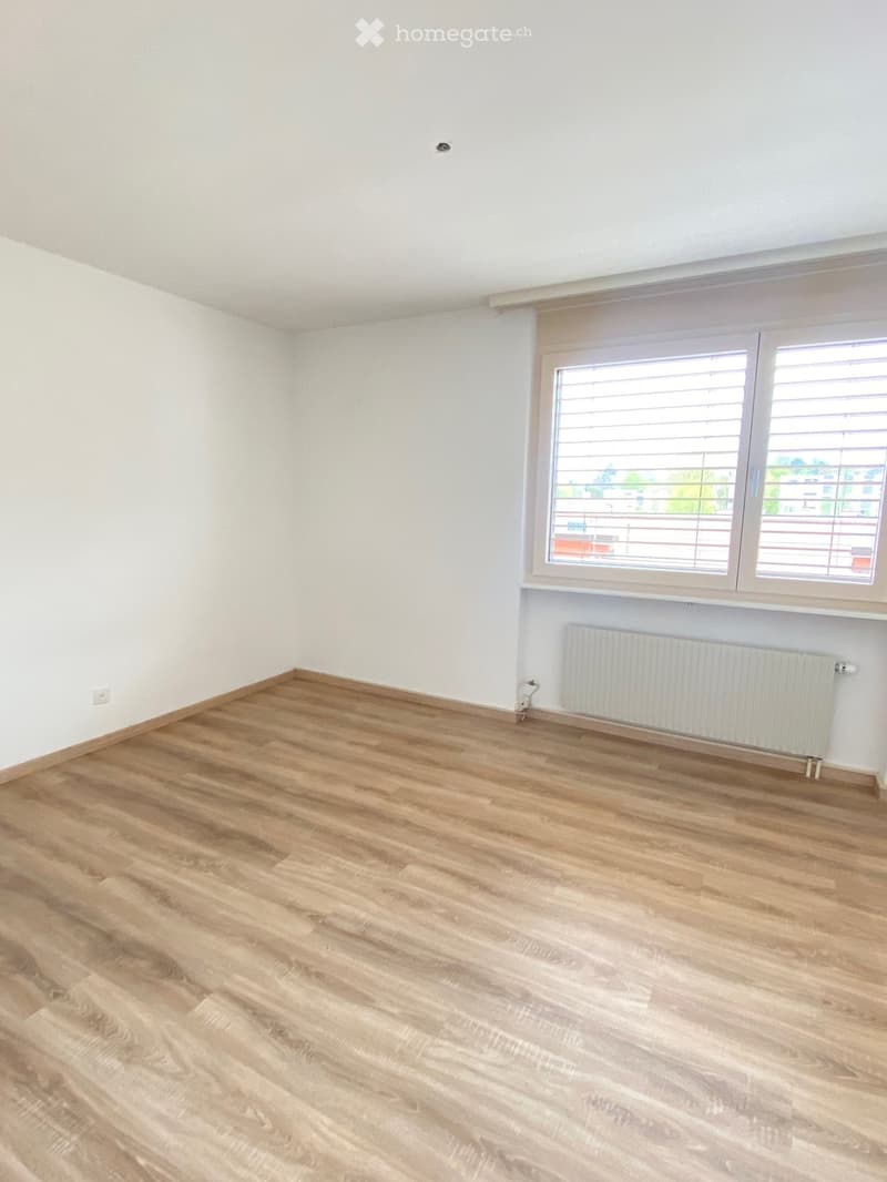 Empty room with light wooden flooring, white walls, and a large window with blinds. A radiator is visible below the window.