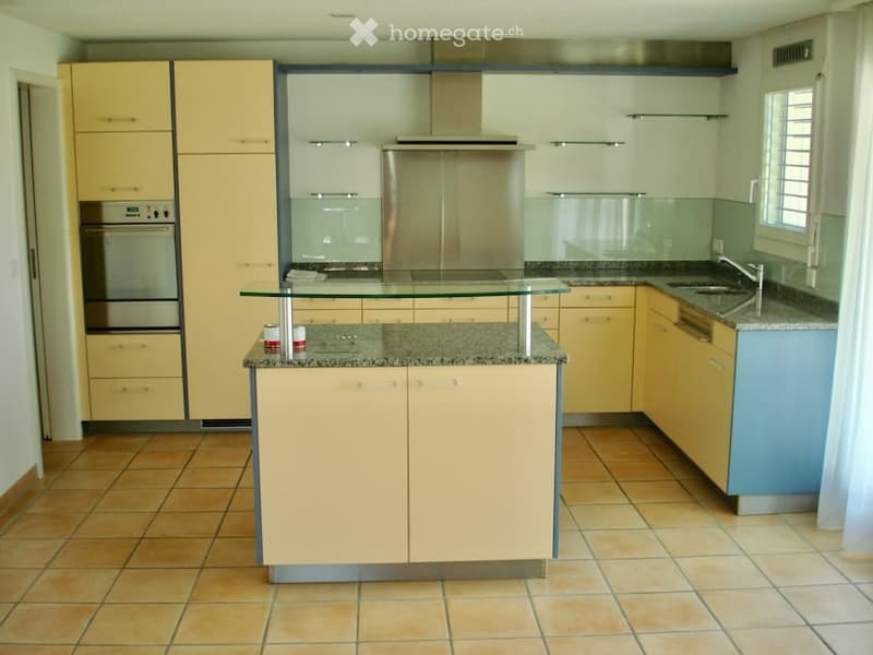 Modern kitchen with light yellow cabinets, granite countertops, stainless steel appliances, and a central island with glass top.