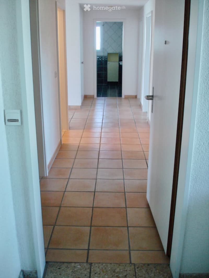 A long, narrow hallway with tiled flooring and white walls, leading to an open doorway at the end.
