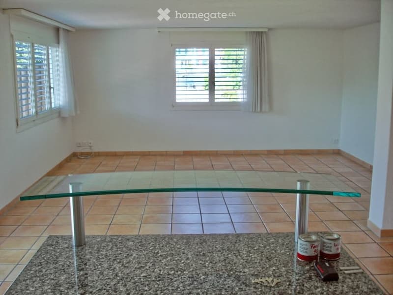 Empty room with tiled floor, large glass table, and a few items on the floor including cans, keys, and a remote control.