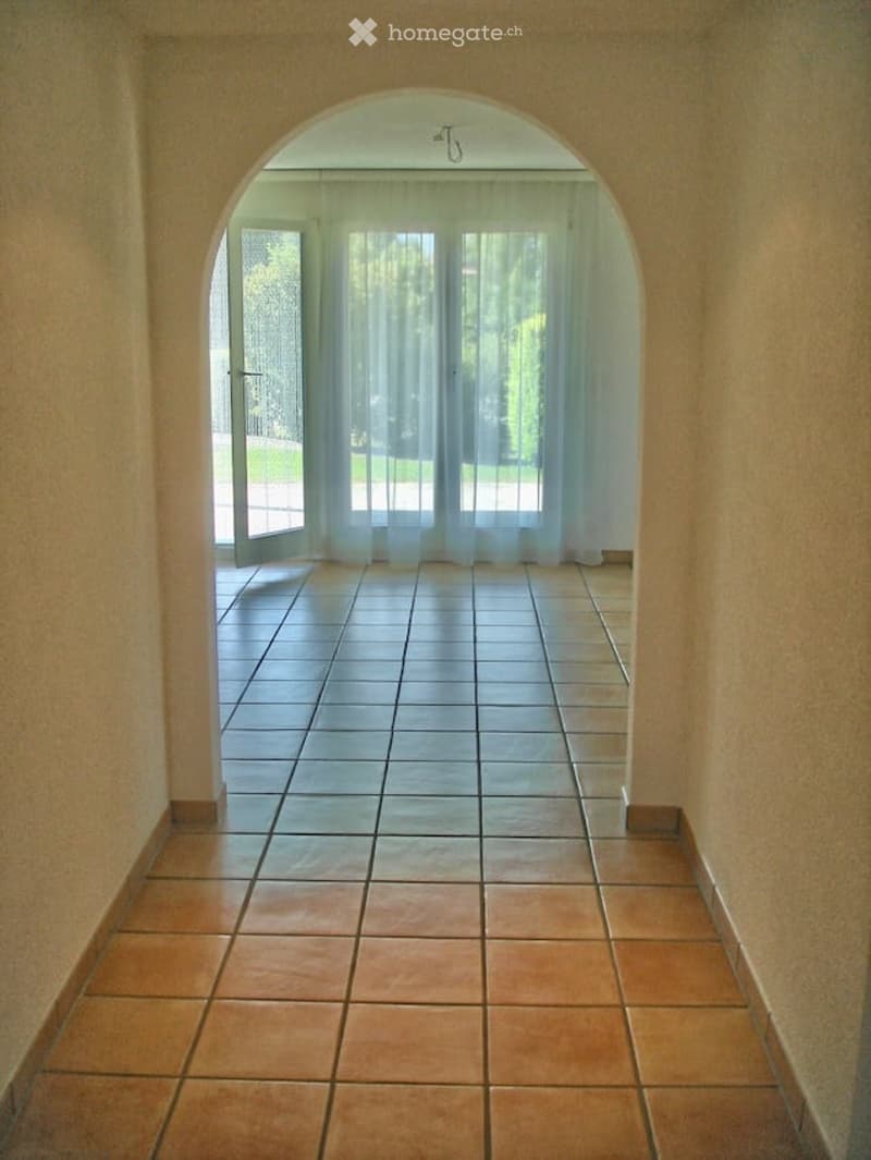 A tiled hallway with an arched doorway leading to a room with large windows and sheer curtains. The floor is tiled in two different colors, and the walls are painted a light beige.