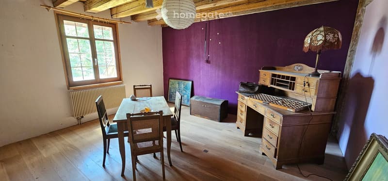 A cozy room with wooden flooring, purple accent wall, and exposed wooden beams. Features a dining table with chairs, a wooden desk with a lamp, and a picture frame on the floor.