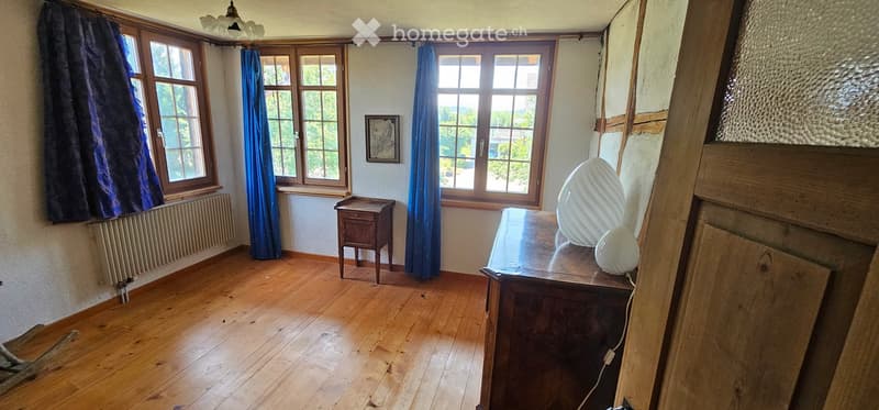 A wooden-floored room with large windows, blue curtains, a small wooden table, a chair, and a decorative white egg-shaped object on a wooden cabinet.