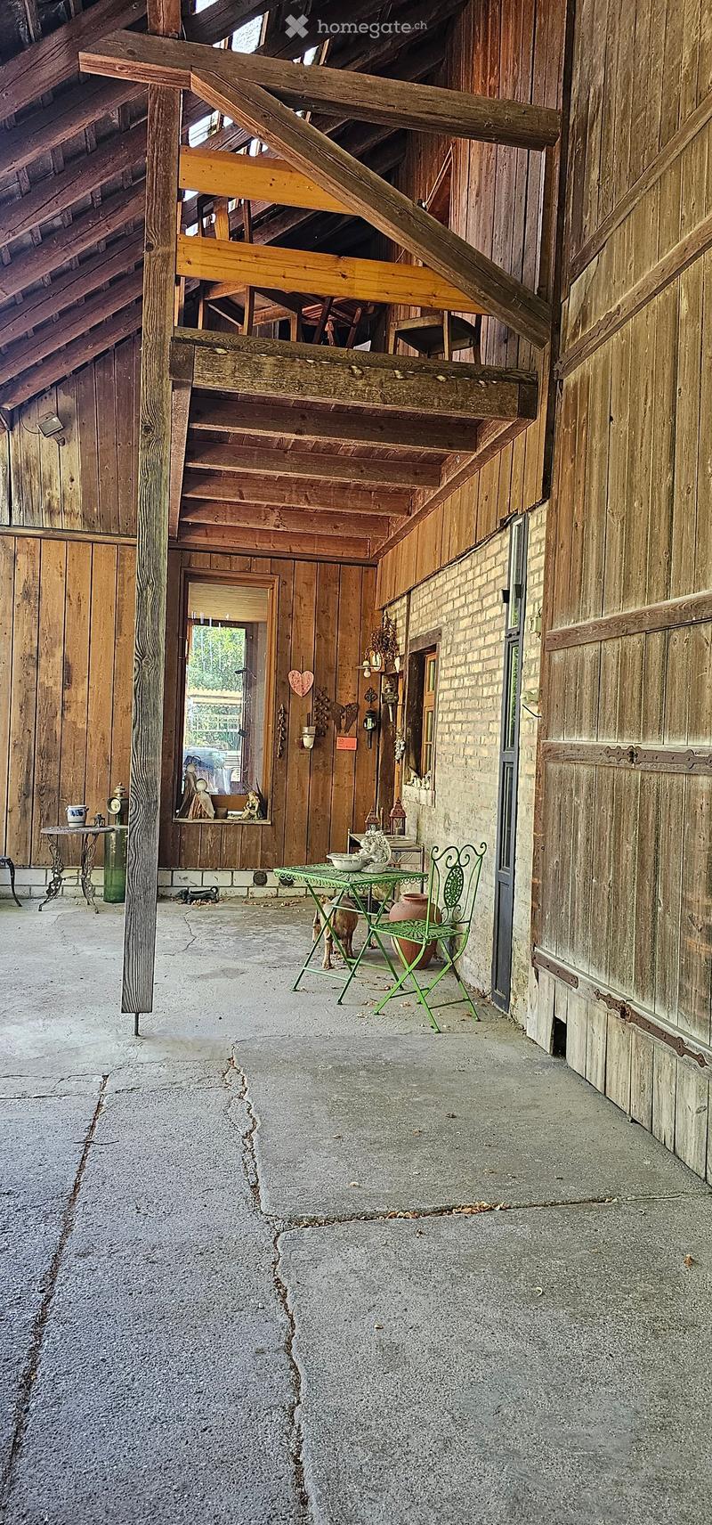 A rustic wooden corridor with exposed beams, wooden walls, and a concrete floor. Features include a green metal table and chair, a small table with a vase, and various decorative items on shelves.
