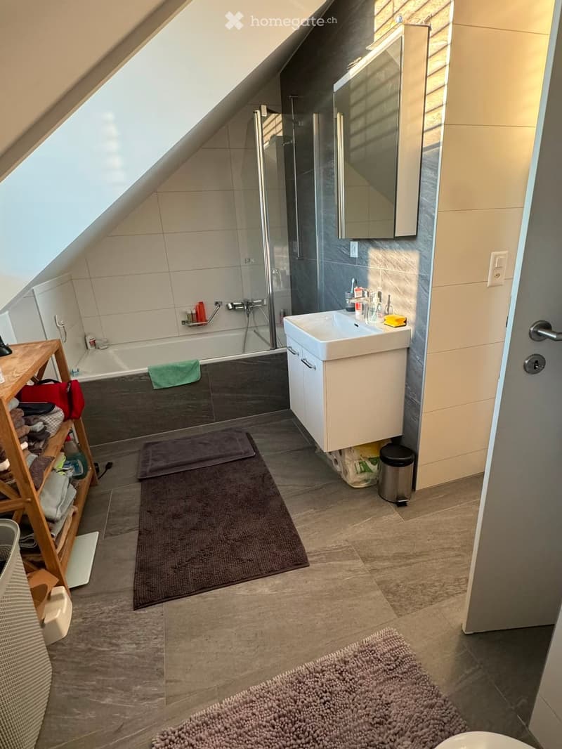 Modern bathroom with a skylight, white sink, bathtub, mirror, and wooden shelf holding towels and accessories.