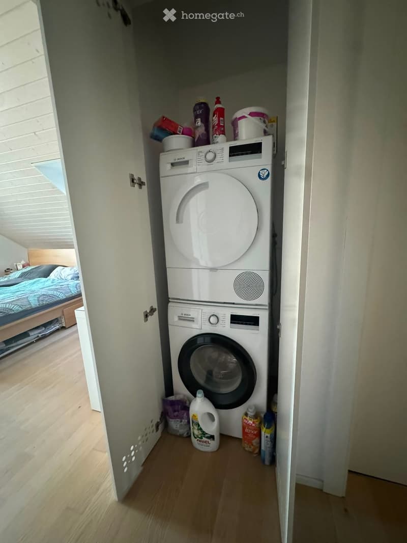 A small, compact laundry area with a stacked washing machine and dishwasher, surrounded by cleaning supplies and a glimpse of a bedroom with a bed and blue bedding.