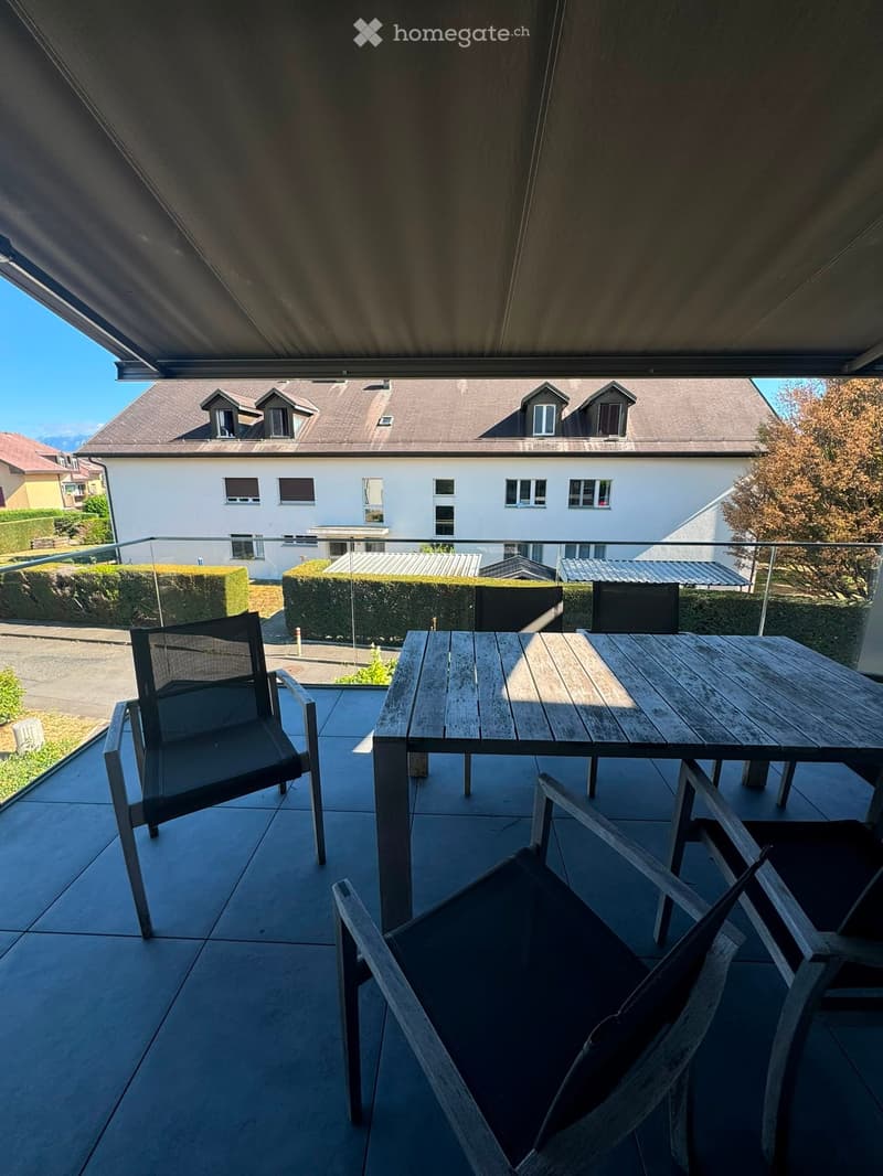 Balcony with a wooden table and two chairs, overlooking a residential area with houses and greenery.