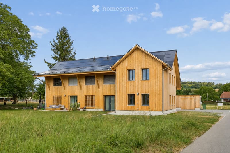 Modern two-story wooden house with solar panels on the roof, large windows, and a spacious front yard with a small seating area.