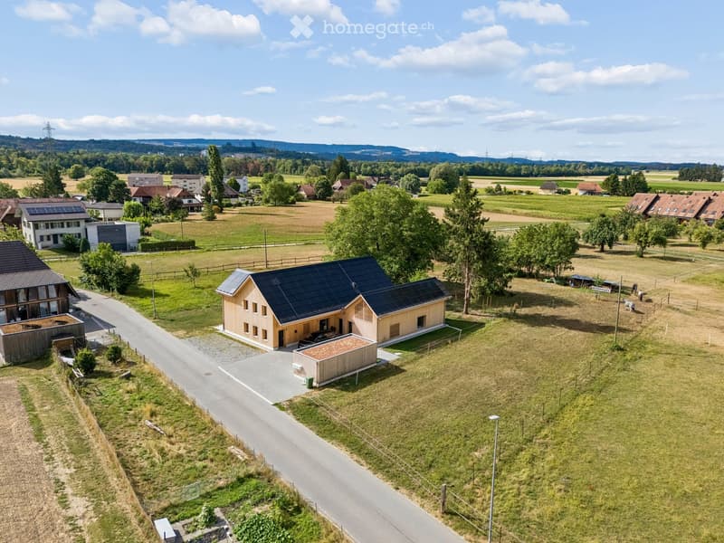 Aerial view of a modern, two-story house with a dark roof and solar panels, surrounded by a rural landscape with fields, trees, and neighboring houses.