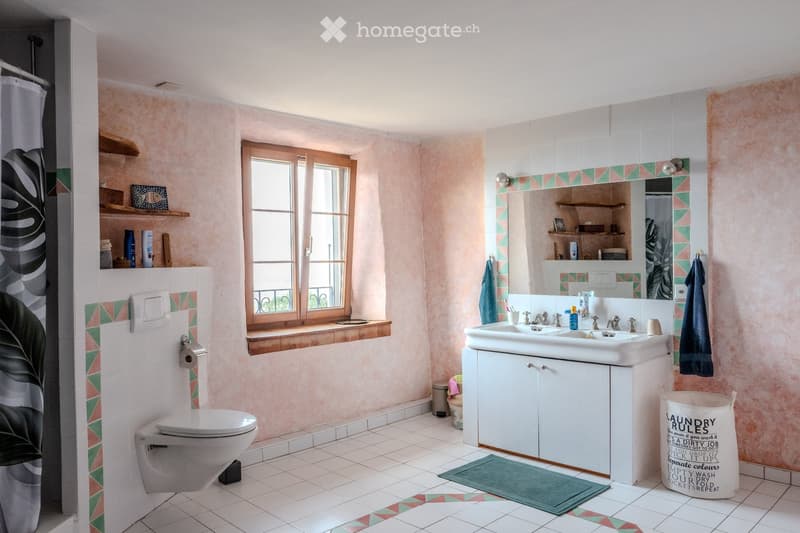 Bright bathroom with pink walls, white tiles, and a large window. Features a toilet, sink with a mirror, cabinet, shelves, and a shower curtain.