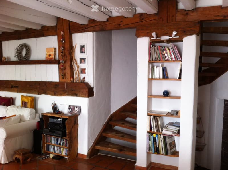 Cozy living room with exposed wooden beams, a staircase leading upwards, a white sofa, a wooden table with various items, and built-in wooden shelves filled with books and decorative objects.