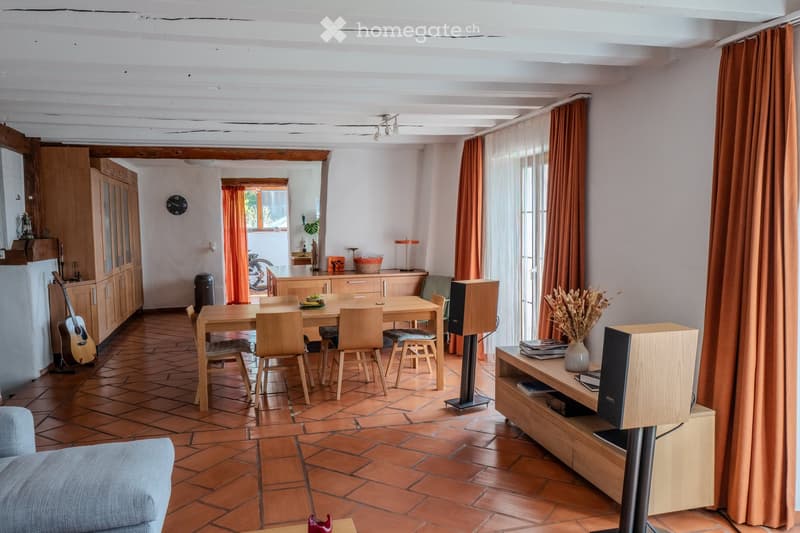 Open-plan living and dining area with wooden furniture, tiled floor, exposed wooden beams, and large windows with curtains.