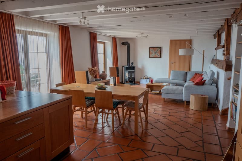 A spacious living room with exposed wooden beams, terracotta tiled floor, and large windows with orange curtains. Features a grey sofa with red cushions, a wooden dining table with chairs, a fireplace, and a floor lamp.