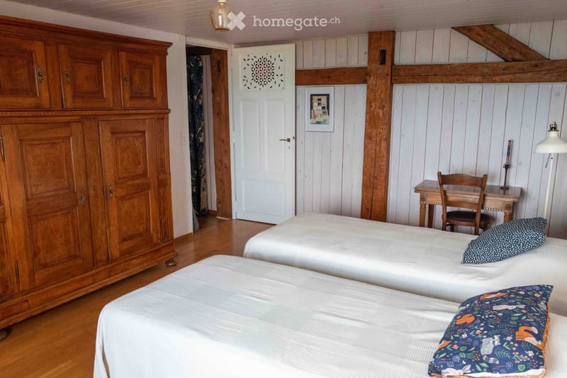 Cozy bedroom with two single beds, a large wooden wardrobe, a small wooden desk with a chair, and a decorative lamp on the wall.