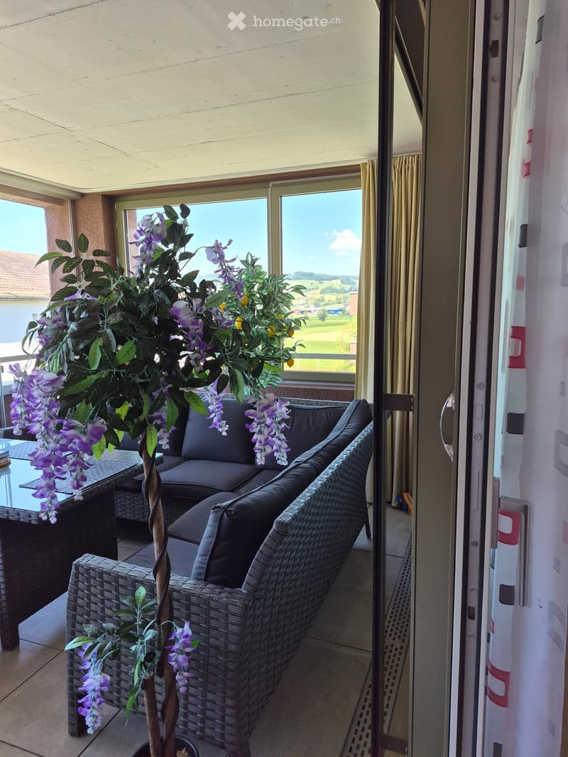 Balcony with a wicker sofa, glass table, and decorative plant with purple flowers, overlooking a scenic countryside view.
