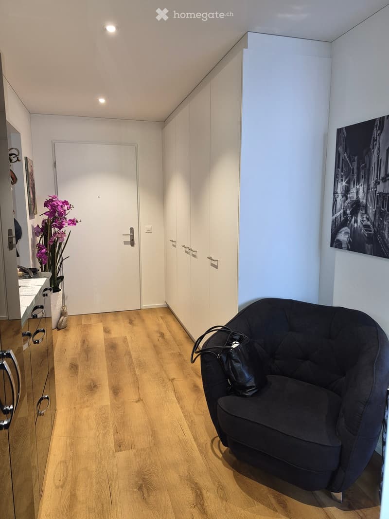 A modern, well-lit hallway with wooden flooring, a black round chair with a handbag, a flower vase with purple flowers, a mirror, and a large picture frame on the wall.