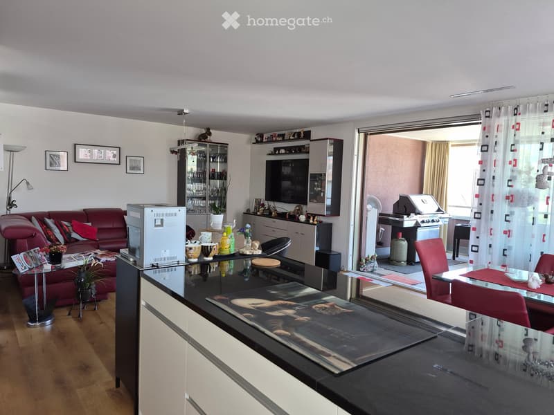Modern living room with a red sofa, glass coffee table, dining area, TV, and bar area. The room features wooden flooring, large windows with curtains, and a balcony area with a grill and outdoor furniture.