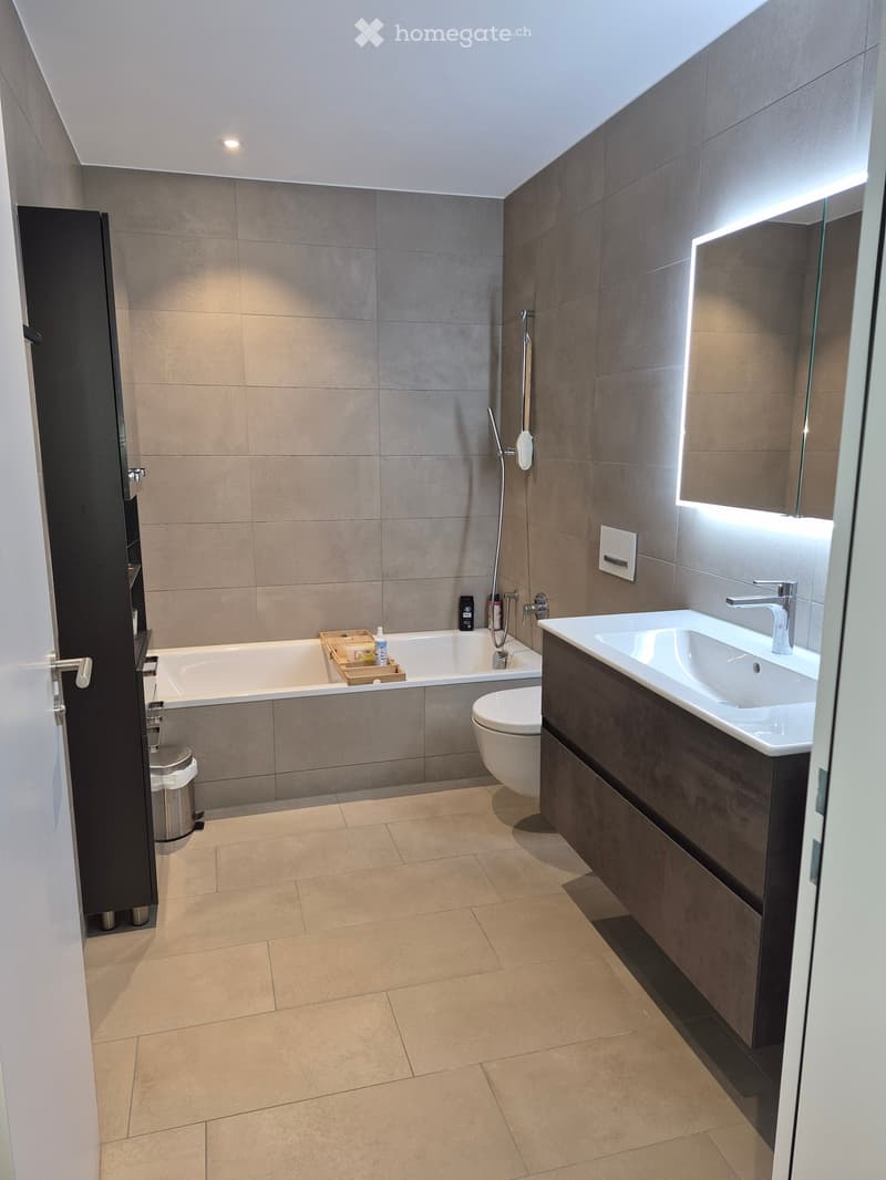 Modern bathroom with a bathtub, toilet, sink, and shower. Features include a large mirror with built-in lighting, a dark wooden vanity, and tiled walls and floor.