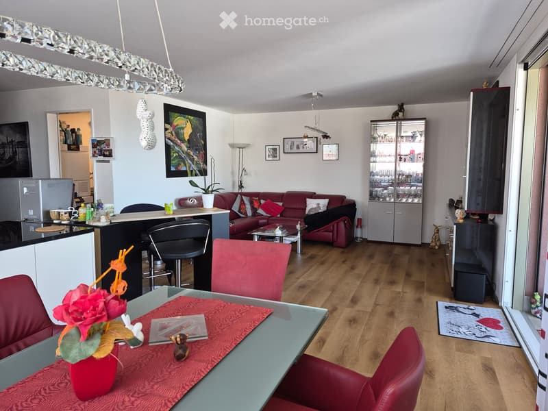 A modern, open-plan living and dining area with a red leather sofa, a glass dining table with red chairs, a kitchen counter with stools, and various decorative items including a flower pot and picture frames.