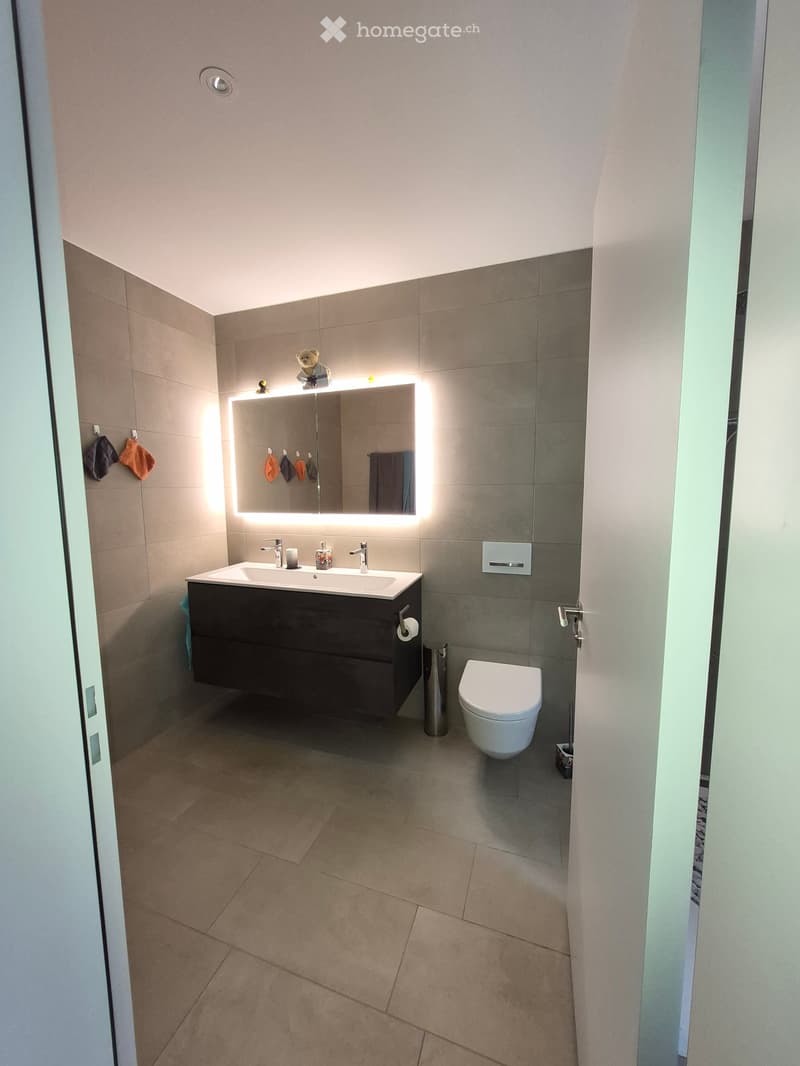 Modern bathroom with a double sink, large illuminated mirror, toilet, and tiled walls and floor.