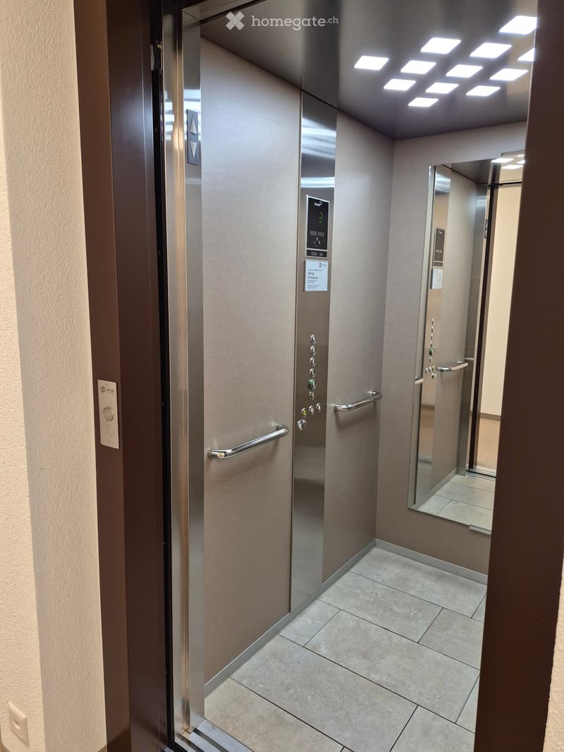 Modern hallway with tiled floor, elevator door, and control panel. The elevator is closed, and the hallway features a clean, contemporary design with neutral tones.