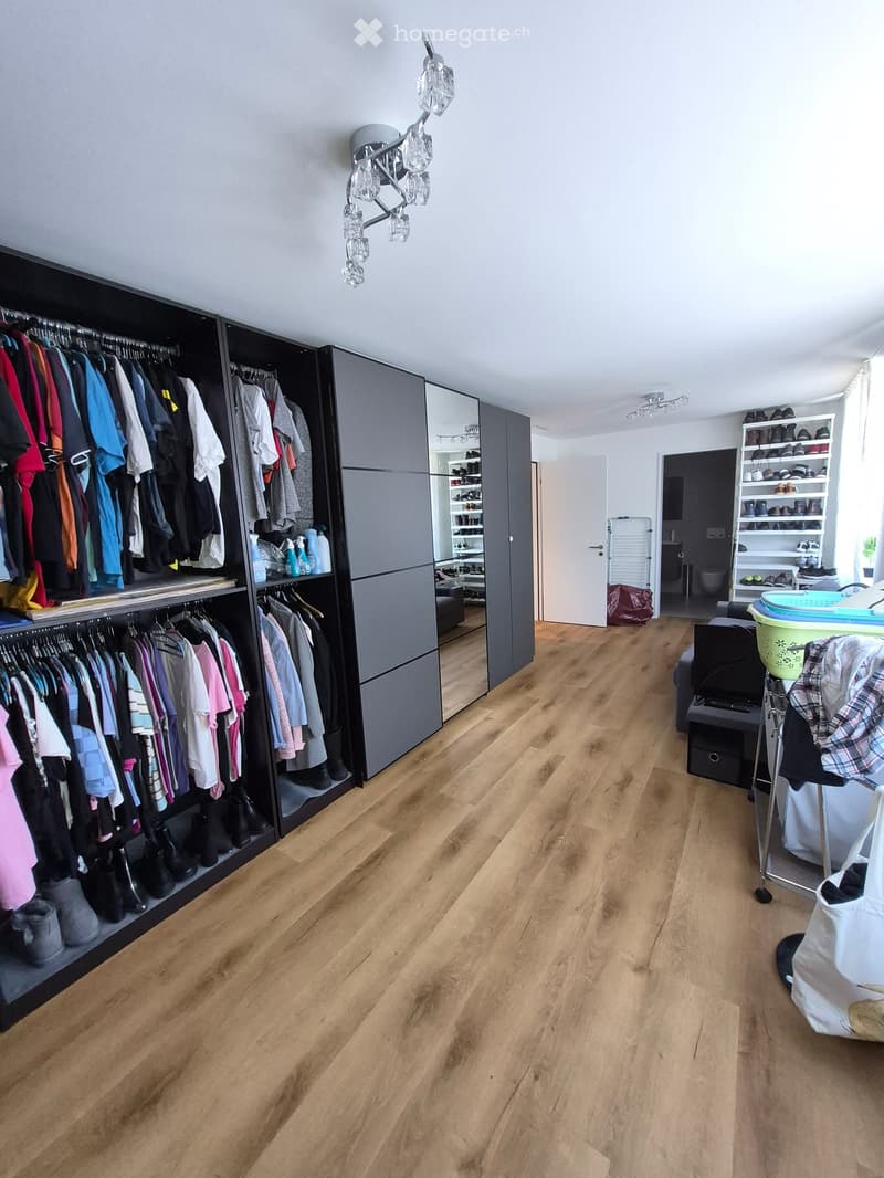 Empty room with wooden flooring, large built-in wardrobe with multiple sections, and a small shelf unit with shoes and other items. The room has modern lighting fixtures and a large mirror on the wardrobe.