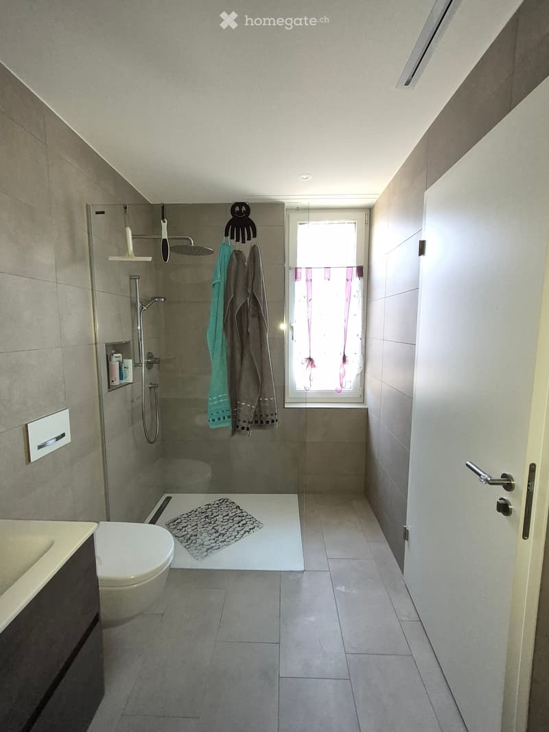 Modern bathroom with a toilet, shower area, towels hanging on a rack, and a window with frosted glass. The floor is tiled, and the walls are clad in light grey tiles.