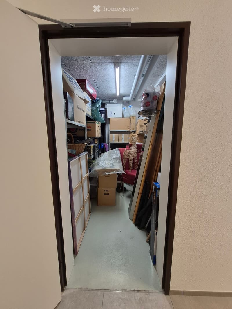 A narrow hallway leading into a storage area filled with boxes, furniture, and miscellaneous items. The hallway has beige walls and a concrete floor.