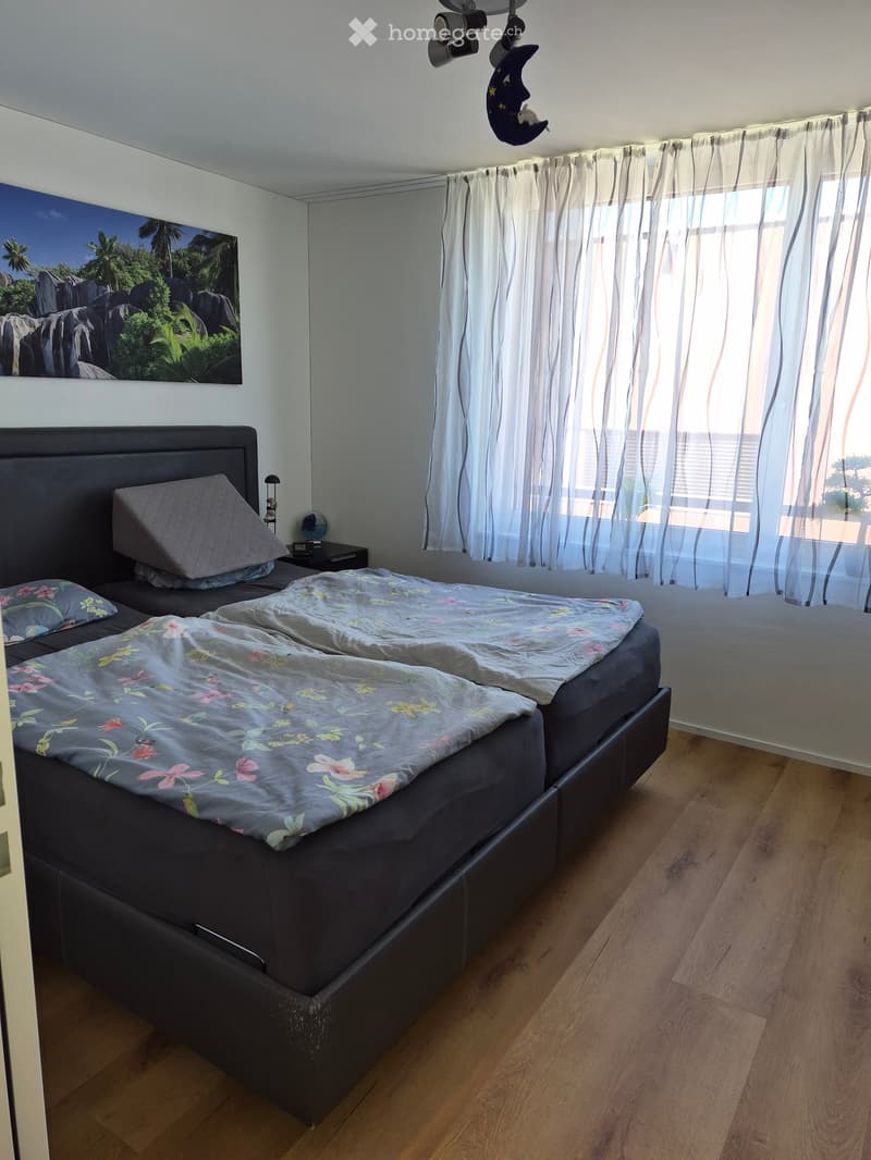 A well-lit bedroom with a large double bed, nightstand, and a picture frame on the wall. The room features wooden flooring and sheer white curtains on the windows.
