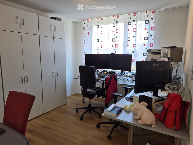 A well-lit room with a wooden floor, a large window with patterned curtains, a desk with two monitors, a chair, a backpack, a stuffed toy, and books.