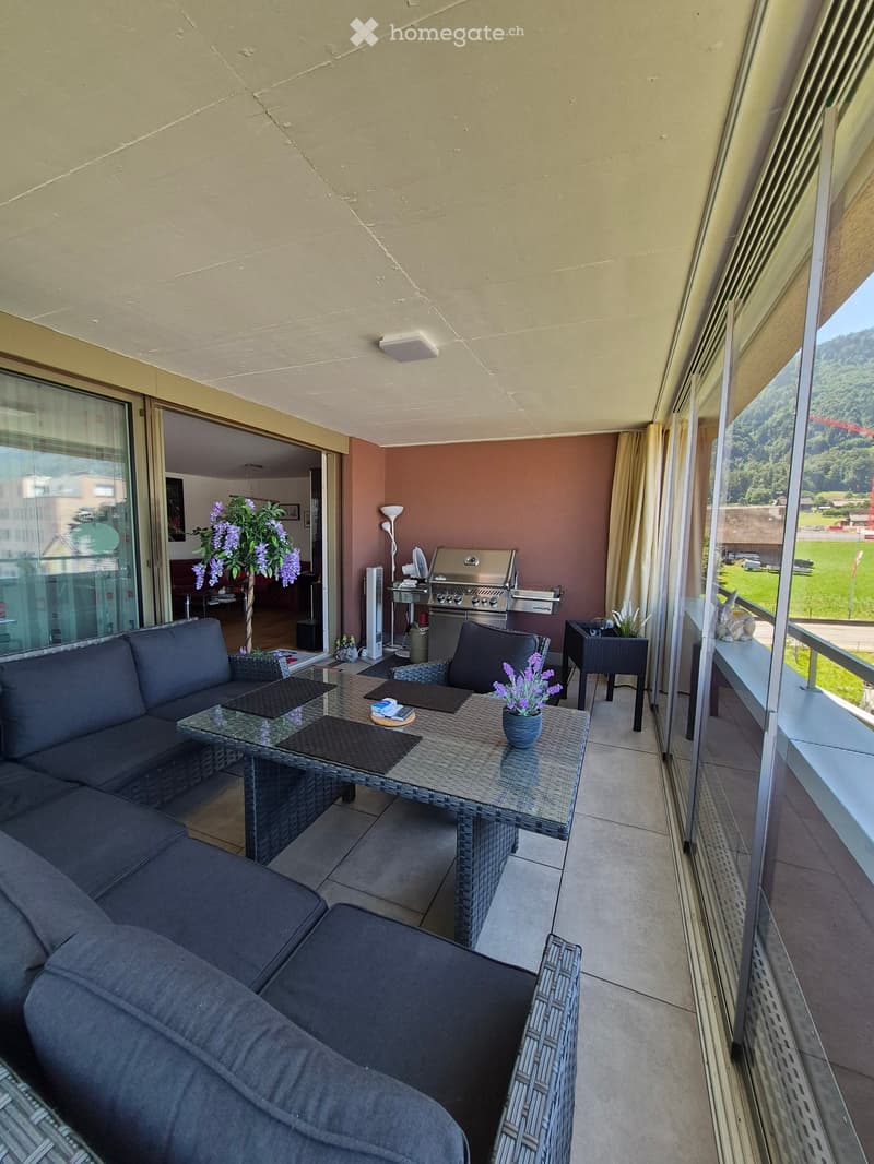 Balcony with a grey sofa, wicker table, chairs, flower pots, and a barbecue. Large windows offer a scenic view of the surrounding landscape.
