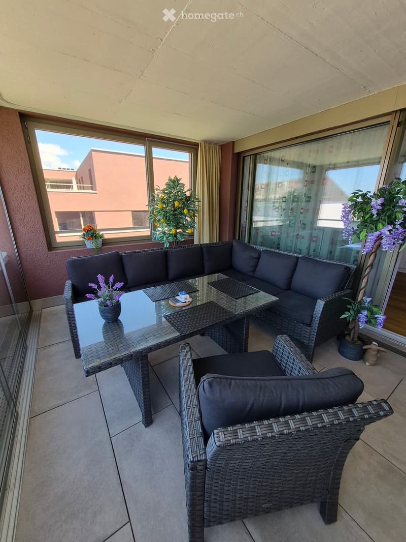 Balcony with a wicker sofa, glass table, two chairs, and flower pots, overlooking a building.