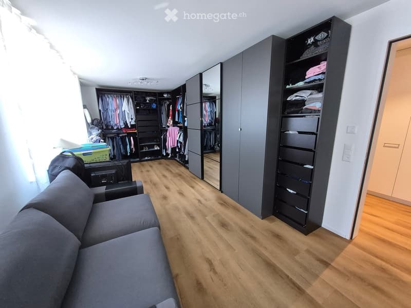 A modern, well-lit room with wooden flooring, a grey sofa, a large wardrobe with mirrored doors, and several storage drawers.