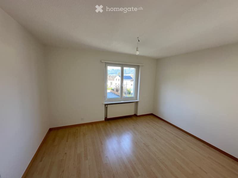 Empty room with wooden flooring, white walls, and a window offering a view of the outside. There is a radiator below the window and a light fixture on the ceiling.