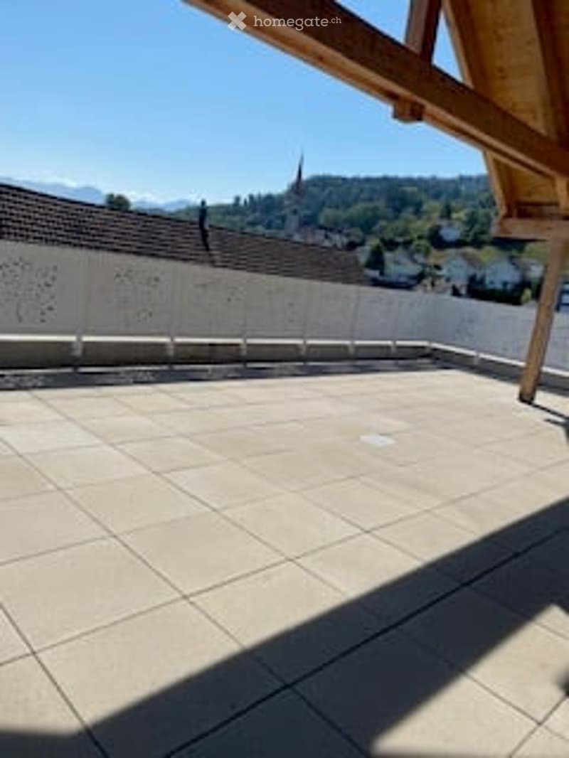A spacious, sunlit balcony with a tiled floor and wooden beams, overlooking a scenic town with visible rooftops and distant mountains.