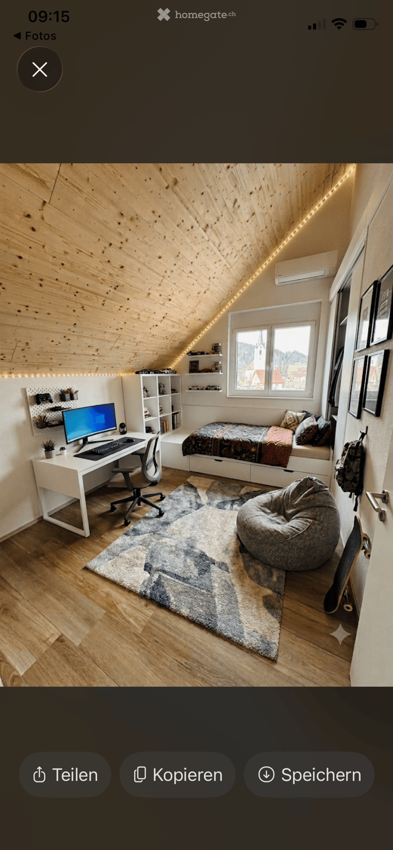 Cozy attic bedroom with wooden walls, slanted ceiling, large window, desk with computer, bed, beanie chair, shelves, and a potted plant.