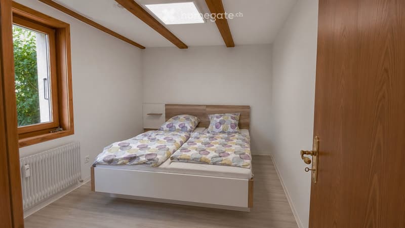 Cozy bedroom with a double bed, matching headboard, patterned pillows, and a radiator under a large window. The room features wooden flooring and exposed wooden beams on the ceiling.