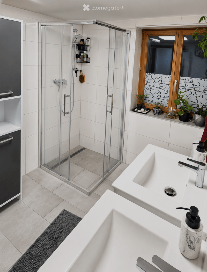 Modern bathroom with white tiles, a large shower enclosure, double sink vanity, bathtub, and decorative plants on the windowsill.
