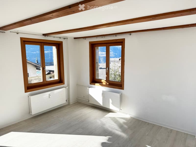 Empty room with wooden flooring, white walls, exposed wooden beams on the ceiling, and two large windows with wooden frames offering a view of the mountains.