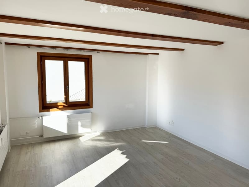 Empty room with light wooden flooring, white walls, and a large window with wooden frame. Exposed wooden beams on the ceiling.
