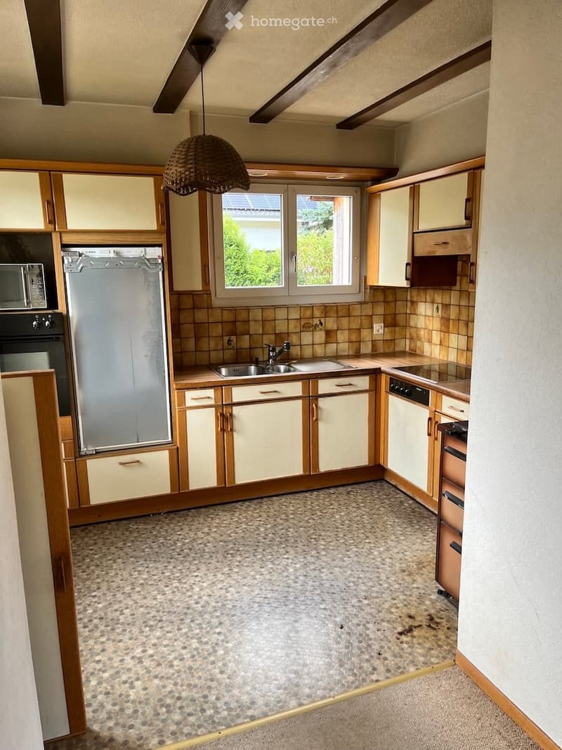 The kitchen features light-colored cabinets with wooden trim, a speckled tile floor, and a window above the sink. Appliances include a refrigerator, oven, microwave, and dishwasher. A hanging woven pendant light is visible.
