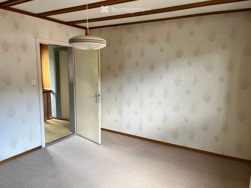 Empty room with light-colored carpet, patterned wallpaper, and a hanging light fixture. The room has a wooden door leading to another space.